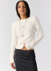 A woman modeling a cream ribbed knit long-sleeve crop sweater with decorative buttons on the cuffs, paired with a black skirt, against a light gray studio background.
