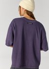 A person shown from the back wearing an oversized purple t-shirt and a cream-colored beanie against a light gray studio background.