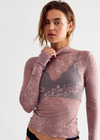 A model wearing a sheer, long-sleeve dusty rose lace top over a gray bra, paired with black lace panties, against a plain light gray background.