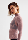 A woman modeling a sheer, long-sleeve mock-neck top in a dusty mauve-pink color with white floral lace applique, paired with black underwear, against a plain light gray studio background.