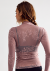 A woman shown from the back wearing a sheer dusty pink long-sleeve top with floral lace patterns over black lace-trimmed underwear, against a plain light gray studio background.