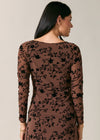 A woman shown from the back wearing a long-sleeve brown dress with a black floral vine pattern, set against a plain light beige studio background.