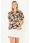 A woman modeling a short-sleeve button-up blouse with a black background and a beige and white floral pattern, paired with a white mini skirt, against a plain white studio background.