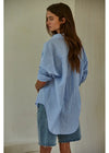 A woman wearing an oversized light blue and white striped button-up shirt with denim shorts, photographed from the back against a plain off-white wall.