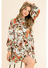 A woman modeling a long-sleeve floral dress with a high neck and gathered waist, featuring an orange, brown, and black floral pattern on a light sage green background, set against a solid beige backdrop.