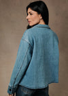 A woman wearing a cropped light blue denim jacket, shown from a three-quarters back view against a textured brown studio backdrop.