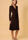 A woman modeling a dark brown long-sleeve wrap dress with a tie waist, standing against a solid beige studio background.