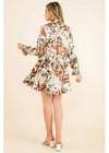 A woman modeling a long-sleeve floral mini dress from the back, featuring a high collar, gathered waist, and a vibrant orange, brown, and green floral pattern on a cream background, set against a solid beige studio backdrop.