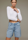 Woman wearing a white long-sleeve top and blue denim shorts against a plain background