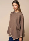 A woman modeling a taupe-brown knit poncho sweater with contrasting cream trim, paired with dark blue jeans, against a solid beige studio background.