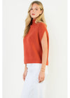 A woman modeling a sleeveless, mock-neck ribbed knit sweater in a warm rust orange color, paired with white pants, against a plain white studio background.