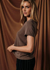 A woman wearing a short-sleeve taupe-brown t-shirt and dark pinstriped trousers, posing in front of a draped brown fabric backdrop.