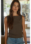 A woman wearing a sleeveless, boat-neck tank top in a solid dark brown color, paired with light-wash blue jeans, standing in a modern indoor setting with a blurred background.