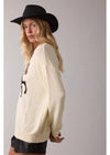 Woman wearing a cream cardigan with a graphic design and a black hat on a plain background
