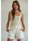 Woman wearing a beige sleeveless top and white shorts against a plain background