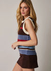 A woman modeling a sleeveless knit sweater vest with horizontal stripes in brown, cream, blue, and red, paired with a dark brown mini skirt, against a plain light gray studio background.