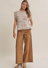A woman modeling a sleeveless knit top with horizontal textured stripes in cream, beige, and brown, paired with wide-leg tan pants and beige ankle boots, against a solid light beige studio background.