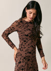 A woman wearing a brown long-sleeve dress with a black floral pattern, posed against a plain beige studio background.