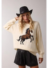 Person wearing a cream sweater with a horse design and a black hat on a plain background