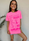 A woman wearing a bright pink graphic t-shirt with 'Monster Mash' printed in pink on the front, posing against a textured white wall.