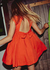 Person wearing an orange dress with a bow detail, standing against a wooden wall.