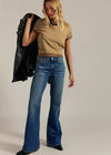 Woman wearing a beige top and blue jeans holding a black jacket against a neutral background