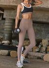 A woman in a gym setting wearing a black and white sports bra and high-waisted taupe leggings, holding dumbbells.