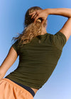 A woman viewed from behind wearing a fitted olive green short-sleeve athletic top and peach-colored shorts against a clear blue sky.