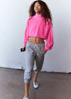 A woman wearing a bright pink cropped sweatshirt and light gray jogger pants with white sneakers, standing against a plain white wall.
