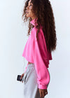 A woman with long, curly brown hair is shown in profile wearing a bright pink cropped sweatshirt and light gray sweatpants against a plain white background.