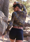 A woman outdoors wearing a fitted long-sleeve crop top with a black, brown, and gold animal print, paired with black athletic shorts.