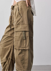 A close-up of a person wearing wide-leg olive cargo pants with multiple flap pockets, side drawstrings, and metal eyelet details, paired with a white lace-trim top against a light gray studio background.
