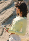 A woman with dark curly hair stands outdoors in a sandy, rocky environment wearing a pale yellow-green long-sleeve ruched crop top and white pants, holding a small booklet.