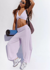 A woman posing in a light purple/lavender activewear set consisting of a twisted bandeau crop top and matching wide-leg sweatpants, holding a blue rolled-up yoga mat and wearing a purple backpack against a white background.