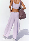 A woman shown from behind wearing a light lavender athletic set consisting of a sports bra and wide-leg sweatpants, carrying a dusty mauve shoulder bag against a white background.