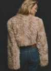 A woman shown from the back wearing a shaggy, cropped faux fur jacket in a light beige color, paired with blue jeans and a brown belt, against a dark background.