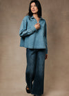 A woman modeling a light blue denim button-up shirt and matching dark blue wide-leg jeans against a textured beige studio backdrop.
