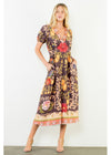 A woman modeling a brown midi dress with a colorful floral and fruit print, featuring puff sleeves, a V-neckline, and a decorative border at the hem, set against a plain white wall.