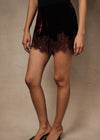A model wearing a short black velvet mini skirt with a dark burgundy lace hem, paired with black pointed-toe mule heels, against a neutral beige studio backdrop.