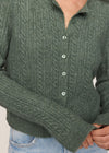 A close-up of a person wearing a textured green cable-knit button-up cardigan sweater with light blue jeans.