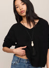 A woman wearing a black knit V-neck sweater with rolled sleeves and a long pendant necklace, paired with blue jeans, against a plain light beige background.