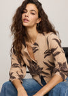 Woman wearing a beige cardigan with leaf pattern and blue jeans sitting on a chair.