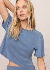 A woman wearing a heathered blue short-sleeve crop top with a side cutout, paired with matching blue bottoms, against a plain beige studio background.