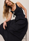 Woman wearing a black dress sitting on a stool against a plain background