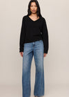 A model stands against a plain beige studio background wearing a black long-sleeve V-neck sweater and blue wide-leg jeans.